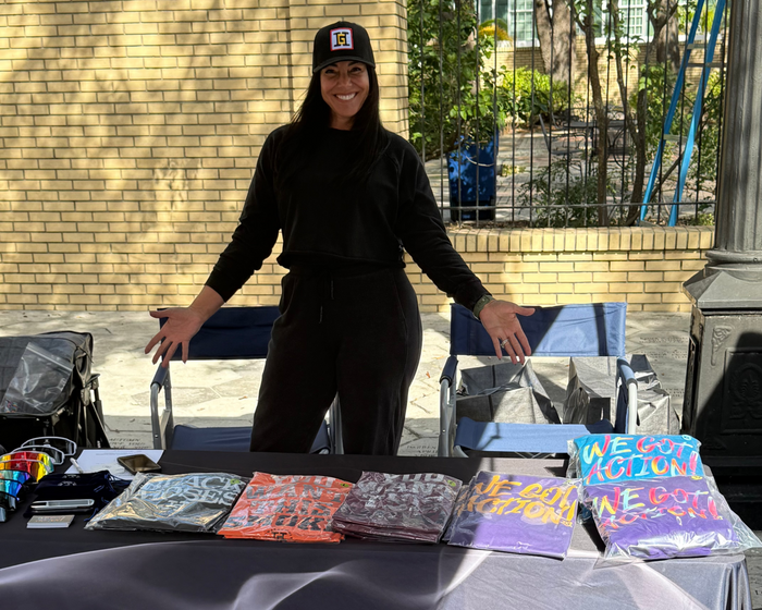Rachel G is standing behind a table with colorful t-shirts and hoodies for sale under a canopy with 'Howard G Cigars' branding.